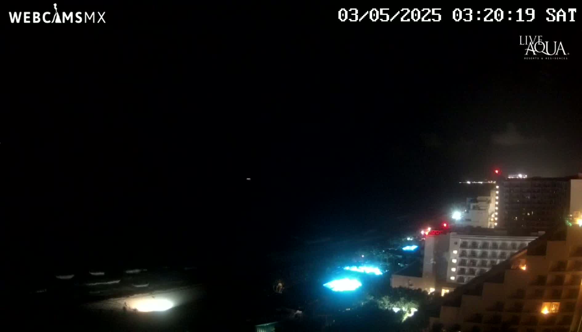 A nighttime view of a beach with dimly lit buildings and a dark ocean under a mostly clear sky.