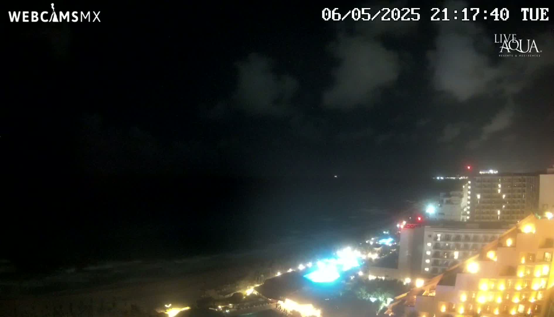 A nighttime view of a coastline with illuminated hotels and a dark, cloudy sky over a gently lapping ocean.
