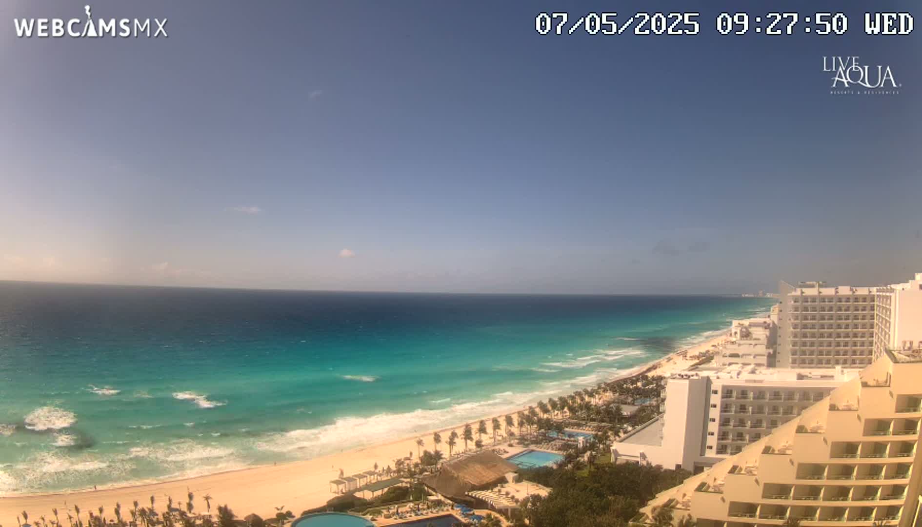 A mostly sunny day shows a long stretch of beach with turquoise water and waves, lined with hotels and palm trees.