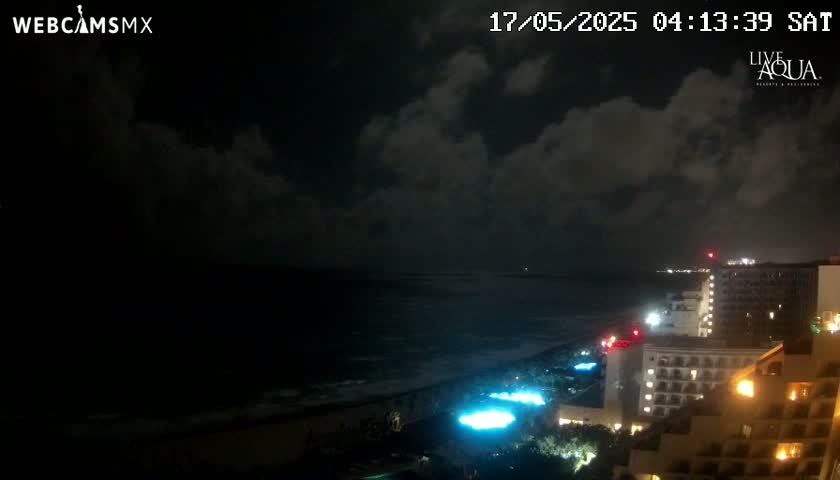A nighttime view of a beach with dark, cloudy skies and illuminated hotels along the shoreline.