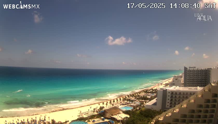 A partly cloudy sunny day shows a long stretch of turquoise ocean meeting a sandy beach lined with hotels and palm trees.