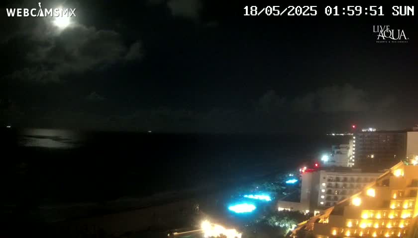 A nighttime view of a moonlit ocean and a brightly lit coastal resort area under a partly cloudy sky.