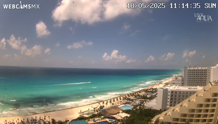 A partly sunny day reveals a beach with turquoise water, waves, and several hotels along the shoreline.
