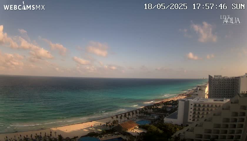 A sunny day with partly cloudy skies overlooks a turquoise ocean meeting a sandy beach lined with hotels and palm trees.