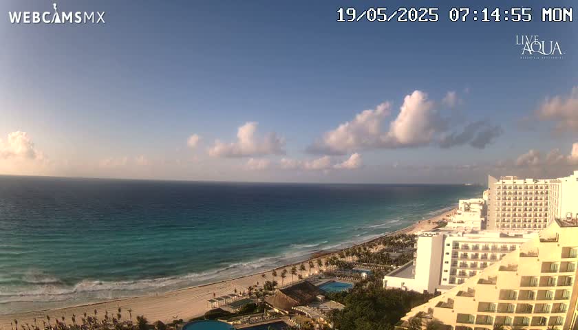 A partly cloudy day reveals a long stretch of sandy beach, turquoise ocean, and several high-rise hotels along the coast.