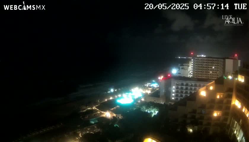 A nighttime view of a coastal resort area with several illuminated high-rise hotels overlooking a dark ocean under a partly cloudy sky.