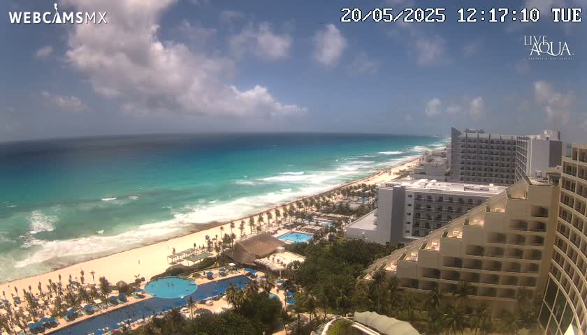 A partly cloudy day shows a beach with turquoise water, waves breaking on the shore, and several high-rise hotels along the coast.