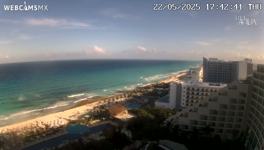 A partly cloudy day shows a panoramic view of a beach with turquoise water, waves, and several resort hotels.