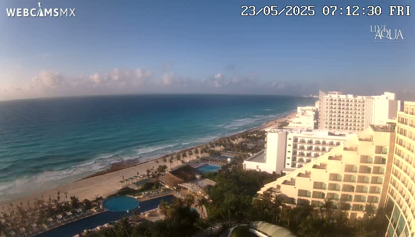 A partly cloudy morning reveals a beach with turquoise water and several hotels along the shoreline.