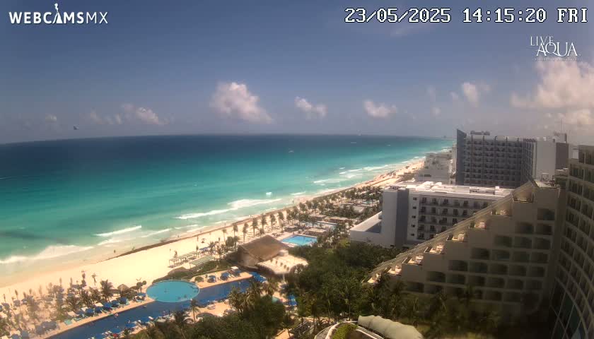A partly cloudy sunny day shows a long stretch of beach and turquoise ocean water next to a row of hotels and resort pools.