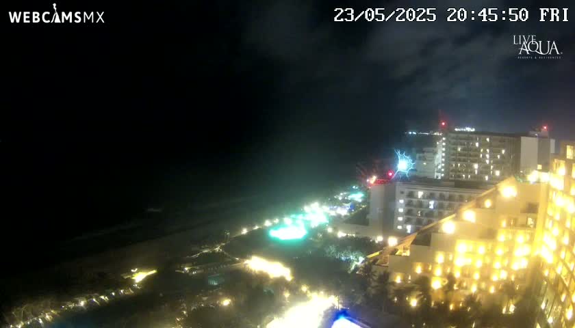 A nighttime, coastal view shows a brightly lit resort complex near a dark ocean under a partly cloudy sky.