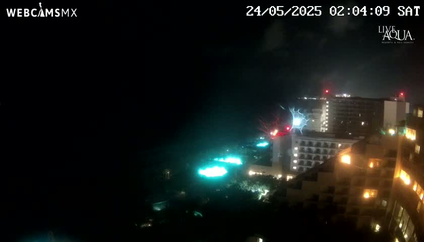 A nighttime view of illuminated buildings and pools, with a lightning strike visible in the distance under a dark, cloudy sky.