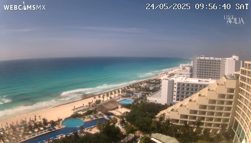 An aerial view shows a tropical beach with turquoise water and waves under a clear blue sky, lined with resort hotels and swimming pools.