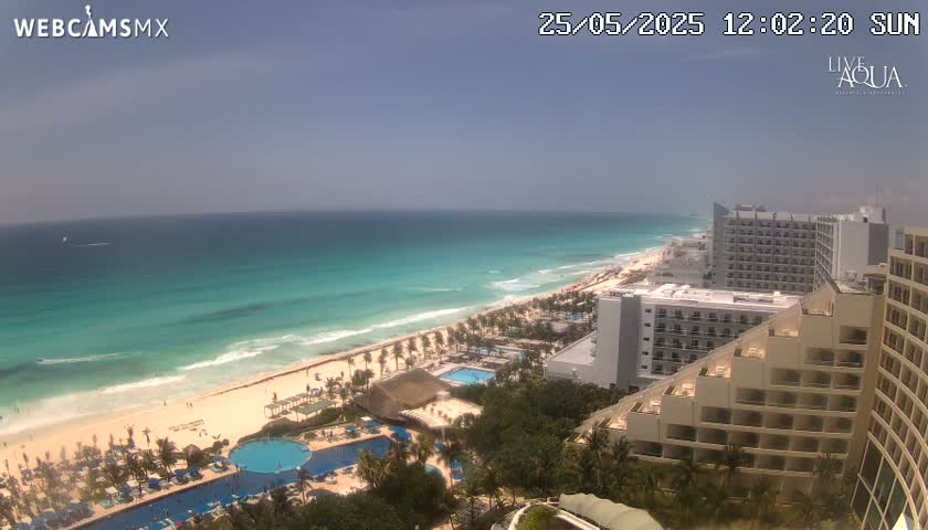 A sunny, aerial view shows a long stretch of turquoise ocean meeting a sandy beach lined with resort hotels and swimming pools.