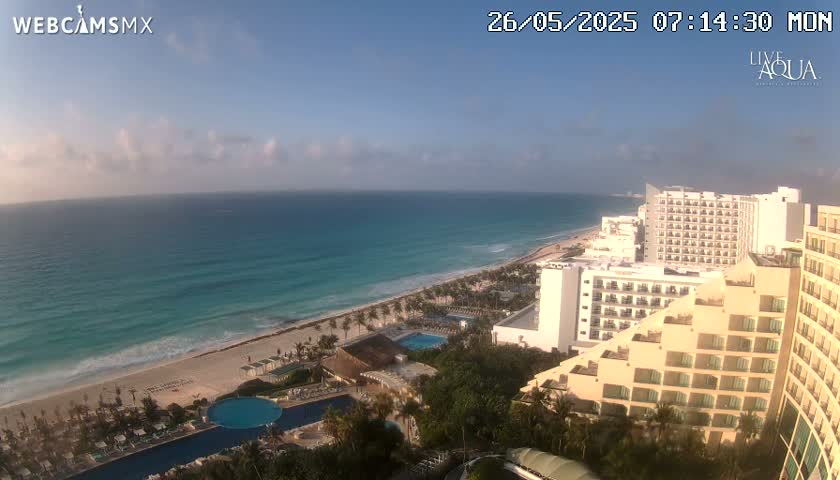 A partly cloudy morning reveals a tropical beach scene with turquoise water, a sandy shore lined with palm trees, and numerous resort hotels.
