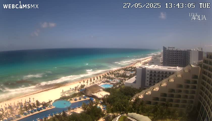 An aerial view shows a sunny beach with turquoise water, waves breaking on the shore, and several high-rise hotels along the coast.