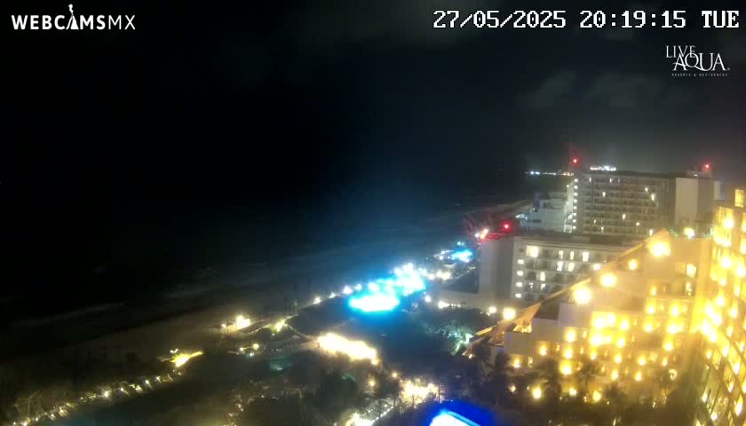 A nighttime, slightly cloudy view of a brightly lit resort complex next to a dark ocean beach.