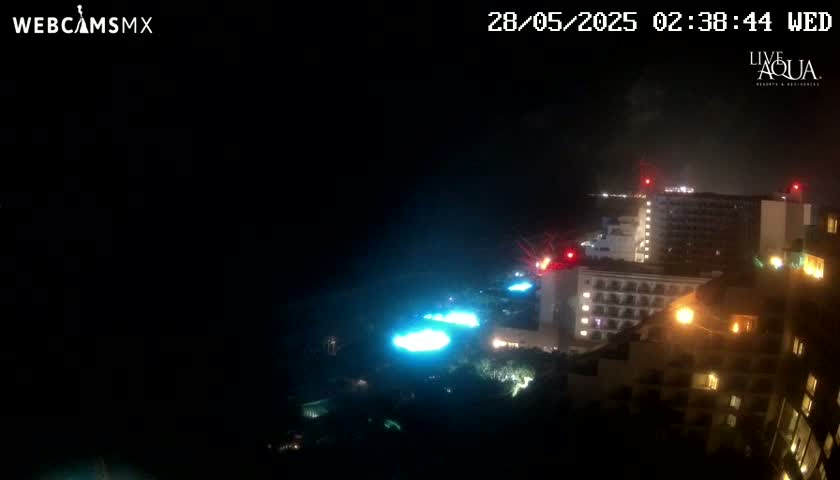 A nighttime view of illuminated hotel buildings and pools, possibly near a coastline, under dark and possibly cloudy conditions.