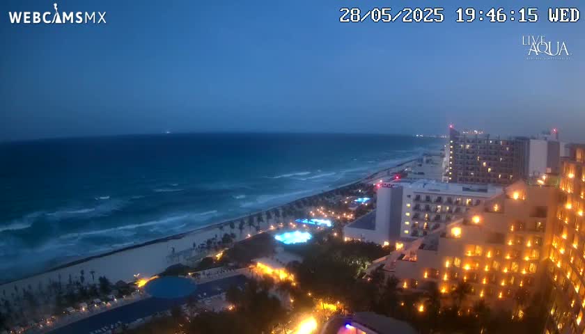 A twilight view of a beach with moderately wavy water and a beachfront resort lit up at night.