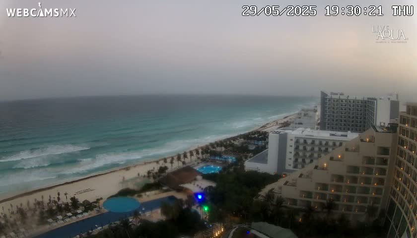 An overcast view of a beach with turquoise water and waves, featuring resort hotels and pools.
