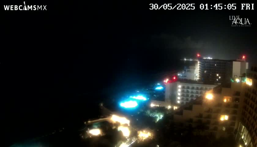 A nighttime view of illuminated hotels and pools near a dark ocean under a mostly clear sky.