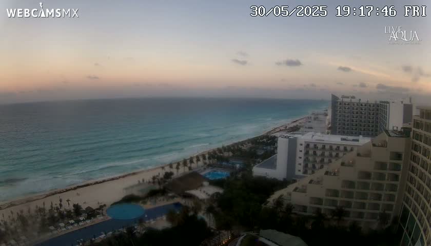 An aerial view at sunset shows a beach with turquoise water, palm trees, and several high-rise hotels.
