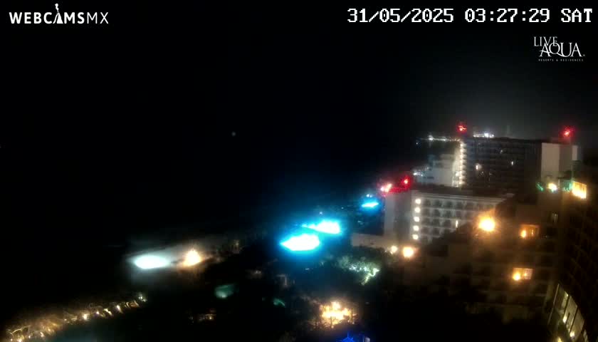 A nighttime view of illuminated buildings near a body of water under hazy conditions.