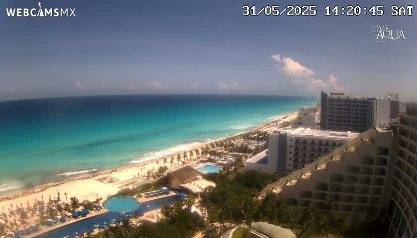 An aerial view shows a beach with turquoise water under a mostly sunny sky, with resort hotels and swimming pools visible along the coast.