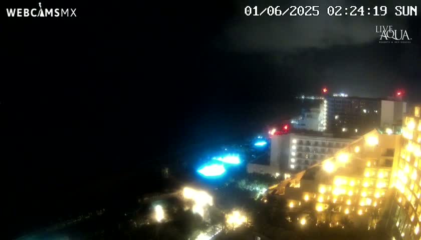 A nighttime, partially cloudy view of illuminated hotels and pools near a dark ocean.