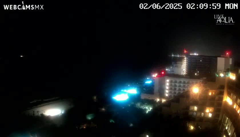 A nighttime, dark, coastal view shows illuminated buildings and pools near a dark ocean under a clear sky.