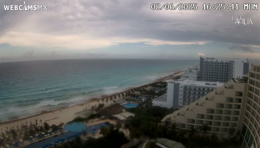 An ocean beach with turquoise water and moderate waves is visible beneath an overcast sky, with several high-rise hotels along the shoreline.