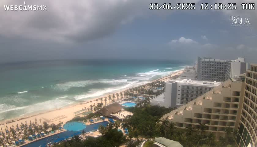 An ocean beach with moderate waves under a partly cloudy sky is viewed from an elevated vantage point overlooking a resort area with several multi-story hotels and swimming pools.