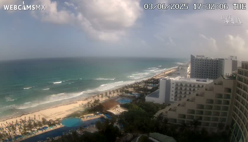 A partly cloudy day reveals a beach with waves breaking along the shore, with resort hotels and swimming pools visible from an elevated vantage point.