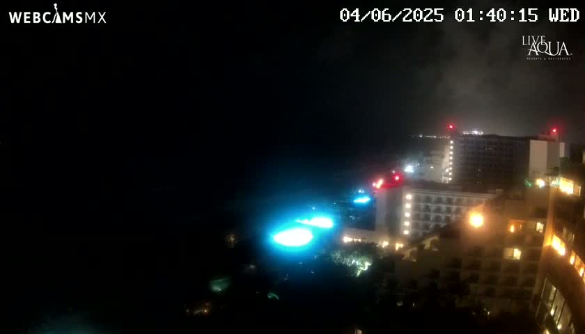 A nighttime, possibly overcast, view of illuminated resort buildings and pools near a dark body of water.
