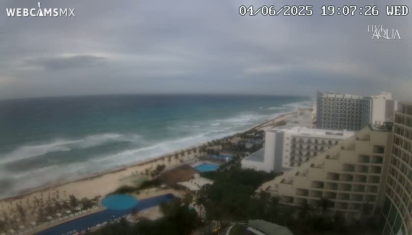 An overcast day shows a beach with moderate waves, hotels, and resort pools.