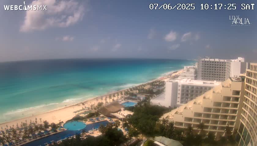 A partly cloudy day shows a long stretch of sandy beach bordering a turquoise ocean, with multiple hotels and resort pools visible in the foreground.
