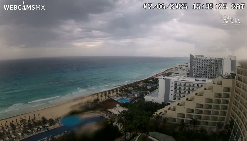 An overcast sky hangs over a beach and resort area with turquoise ocean waves lapping the shore.