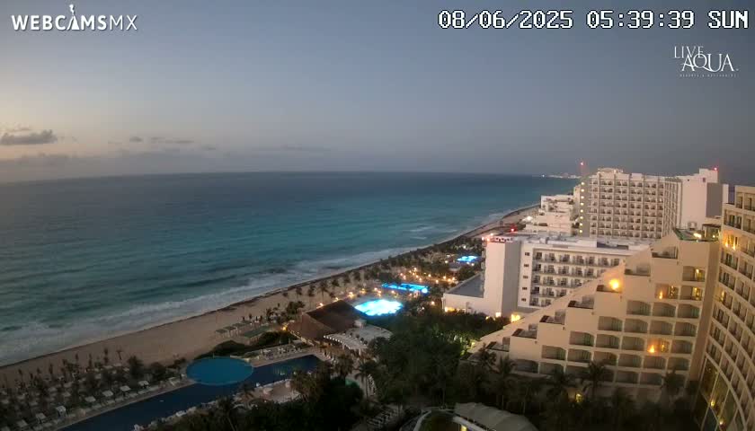 A twilight view of a beach resort at night, showing a sandy beach, ocean waves, and numerous lit hotel buildings.