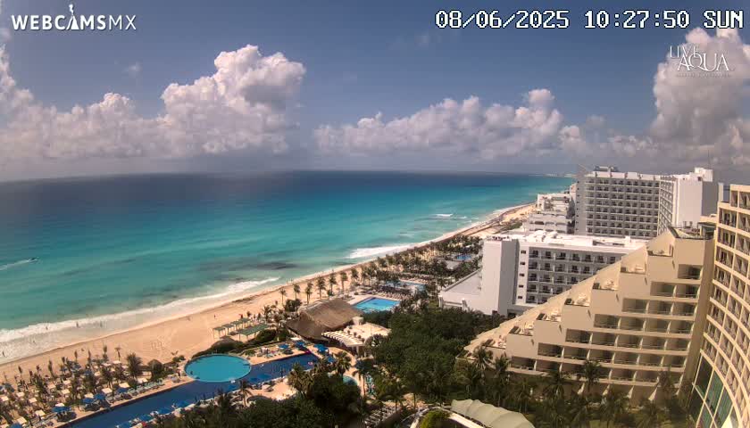 A partly cloudy sunny day overlooks a tropical beach with turquoise water, numerous hotels, and swimming pools.