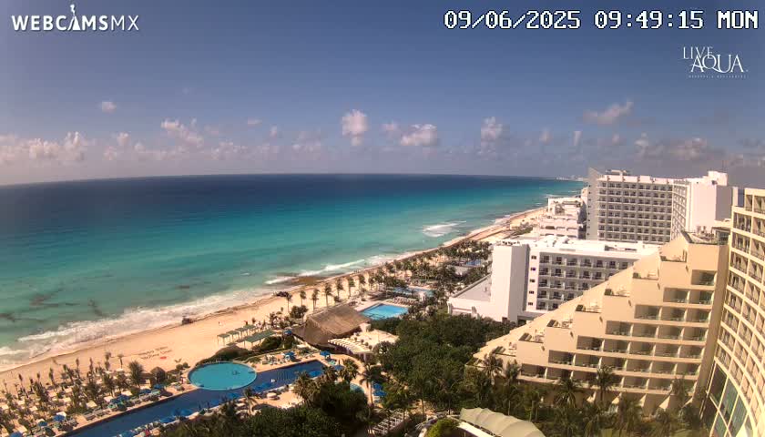 A sunny day shows a beach with turquoise water, lined with hotels and palm trees, and a large swimming pool.