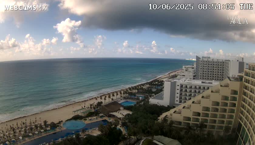 An ocean view from a high vantage point shows a partly cloudy sky, a beach with resorts and pools, and the ocean under partly cloudy skies.