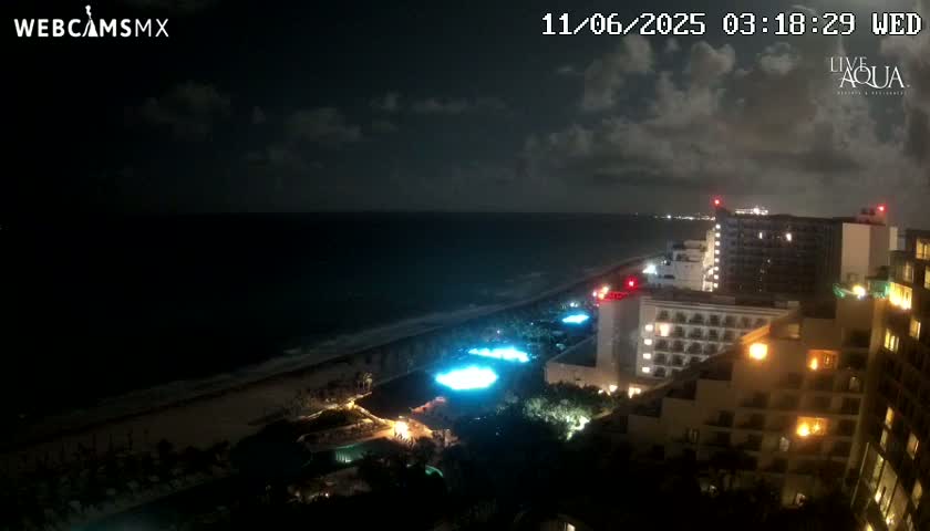 A nighttime view of a coastline shows a dark ocean, a sandy beach, illuminated resort buildings, and a partly cloudy sky.