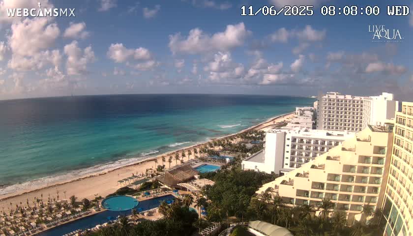 A mostly sunny day reveals a tropical beach with turquoise water, lined with hotels and palm trees.