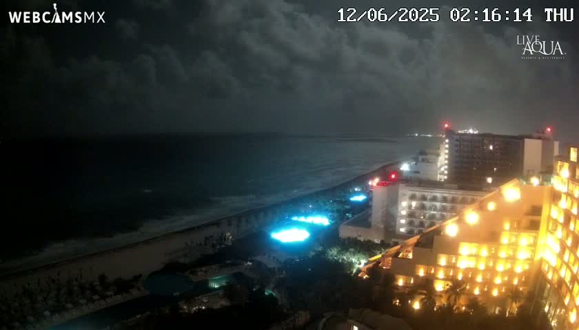 A nighttime coastal view shows a dark ocean under a cloudy sky, with brightly lit hotels and pools along a beach.