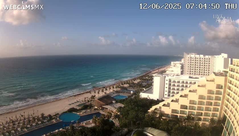 A partly cloudy morning shows a beach with turquoise water and numerous beachfront hotels and resorts.