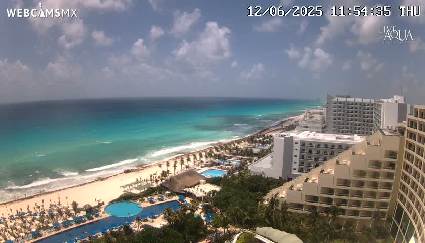 An aerial view shows a beach with turquoise water under a partly cloudy sky, and multiple resort hotels with pools are visible along the coastline.