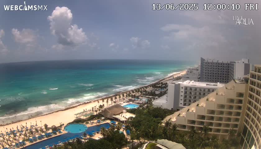 A partly cloudy day shows a beach with turquoise water, waves, and resort hotels, with multiple pools visible near the ocean.
