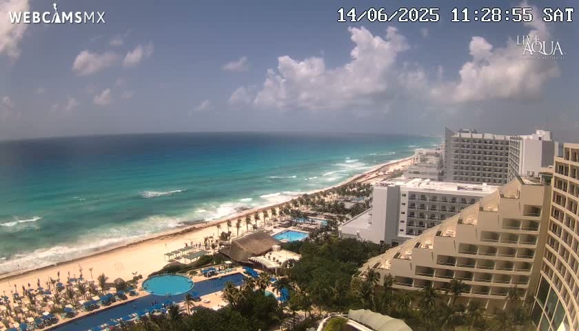A partly cloudy day shows a beach with turquoise water, hotels, and swimming pools.
