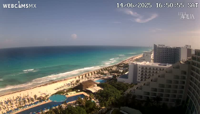 A partly cloudy day shows a beach with turquoise water, hotels, and swimming pools.