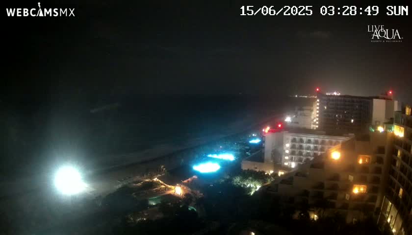 A nighttime coastal view shows brightly lit hotels and pools along a beach under a dark, cloudy sky.